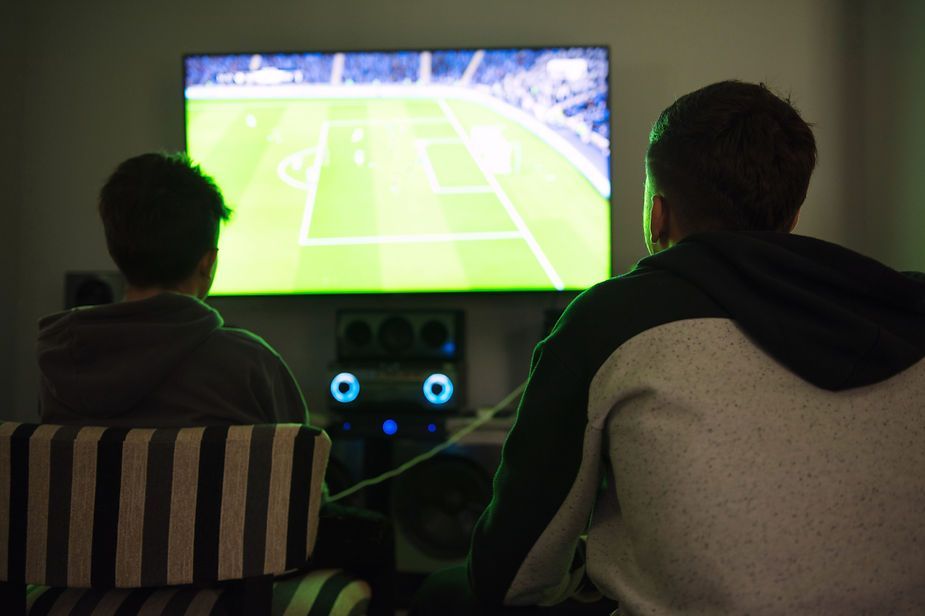 Two men are sitting in front of a television watching a soccer game.