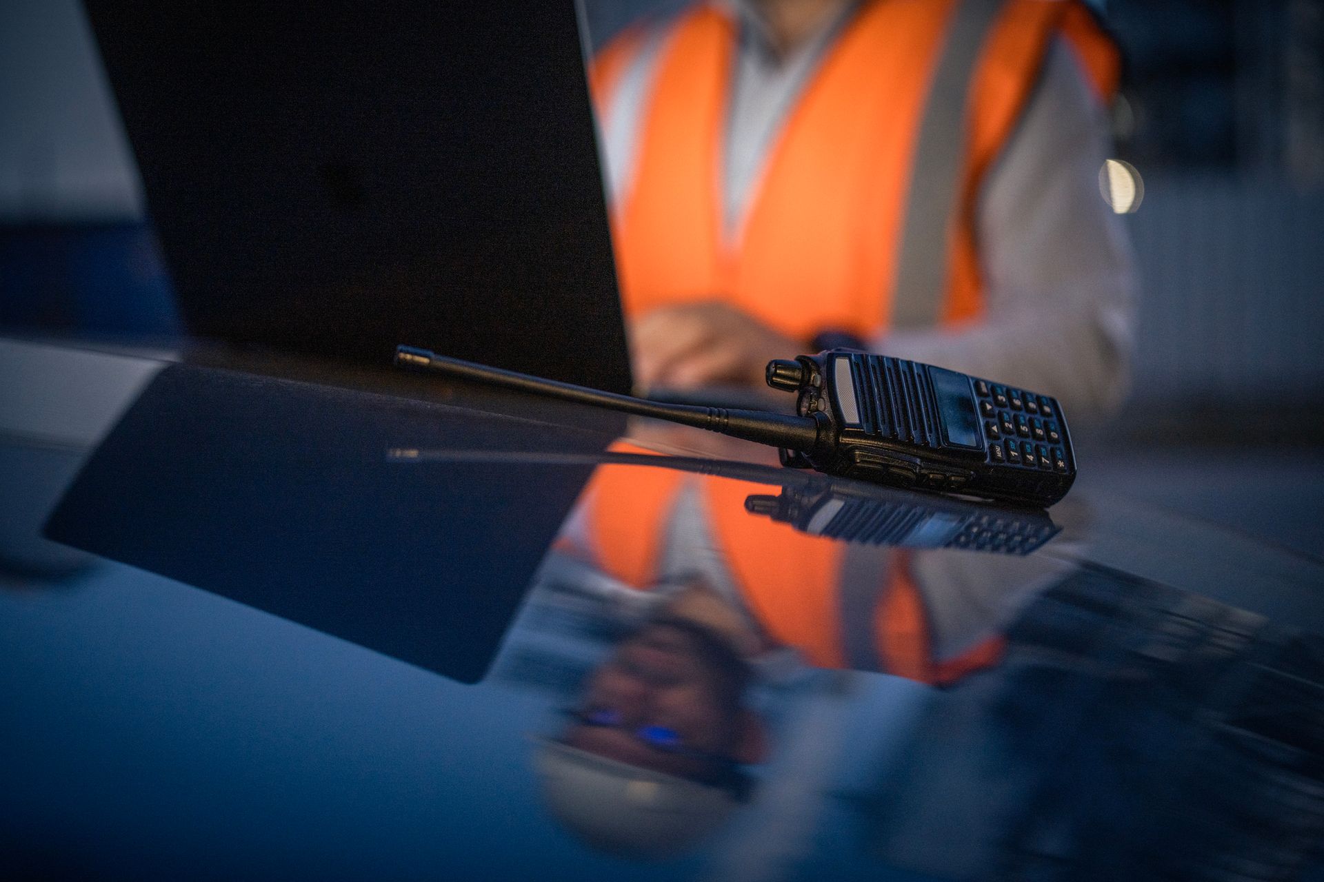 A man in an orange vest is using a laptop and a walkie talkie.