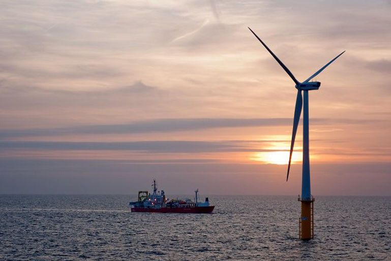 A wind turbine in the middle of the ocean with a boat in the background