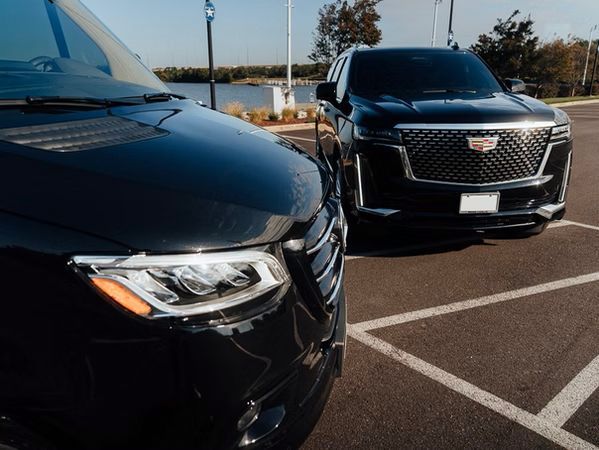 Black vehicles parked in a lot: a van on the left, a Cadillac SUV on the right. Outdoors, sunny day.