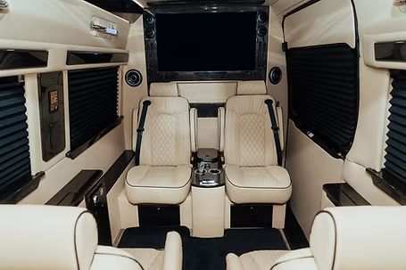 Interior of a luxury van with cream-colored leather seating, a large screen, and dark window shades. Interior of a luxury van with cream-colored leather seating, a large screen, and dark window shades.