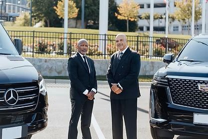 Two Black men in suits stand between a Mercedes van and Cadillac Escalade. Outdoors, sunny setting. Two Black men in suits stand between a Mercedes van and Cadillac Escalade. Outdoors, sunny setting.