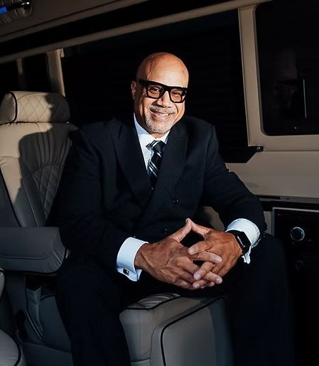 Man in suit, glasses, smiling, sitting in a luxury vehicle.