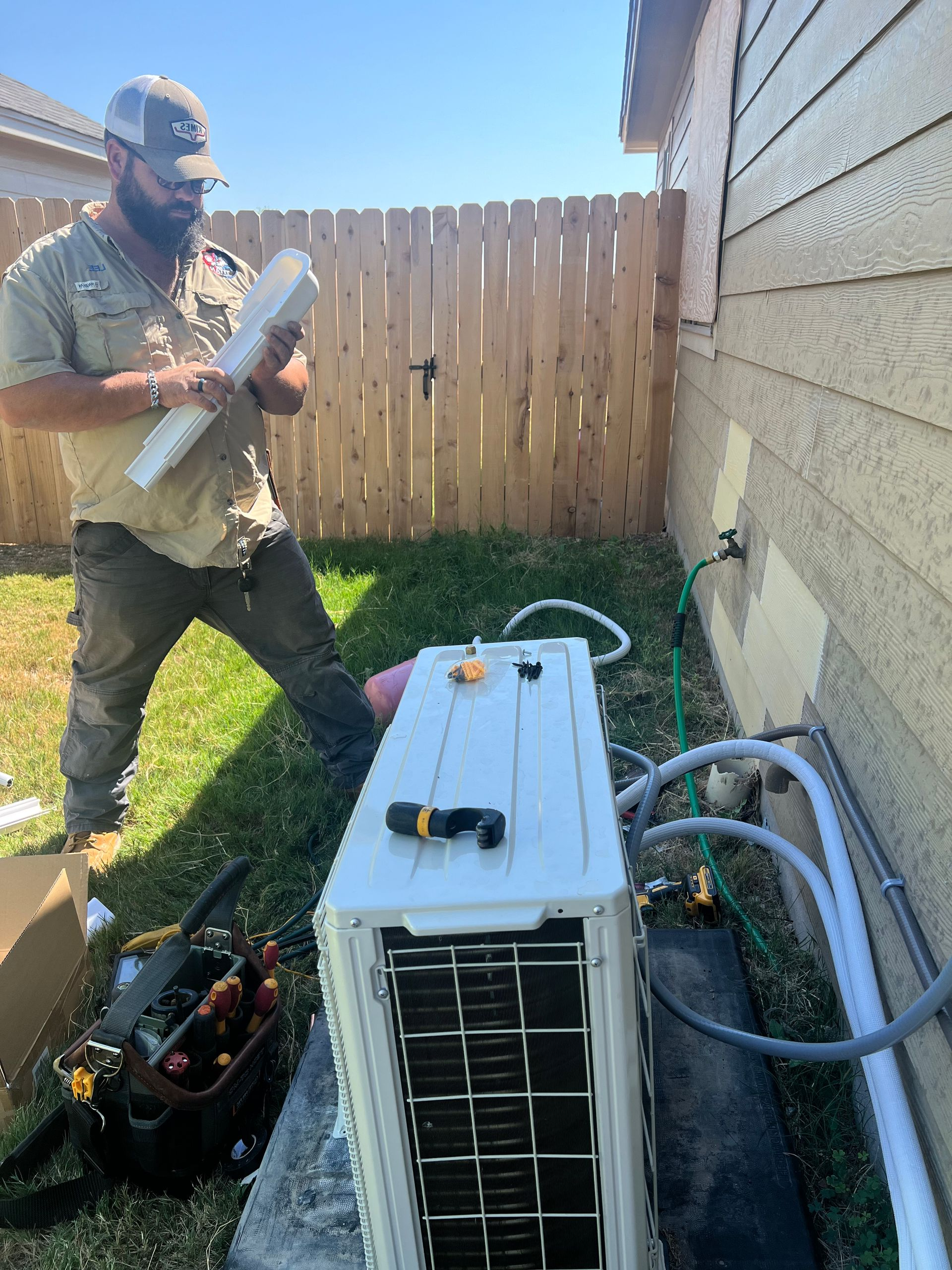 HVAC technician in red shirt adjusts wiring on a white outdoor unit, wearing gloves and safety glasses.