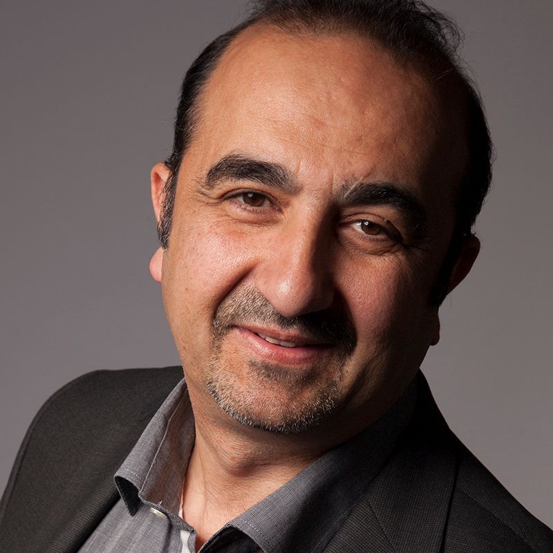 Een man in een grijs shirt en een donkere blazer lacht naar de camera tegen een grijze achtergrond.