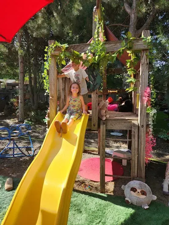 Child on yellow slide, another at top of wooden play structure with faux vines; green grass, red shade.