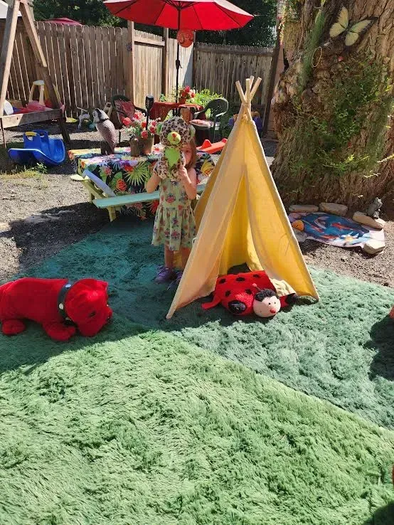 Girl playing with toys near a tent and red umbrella in a backyard with green rug.