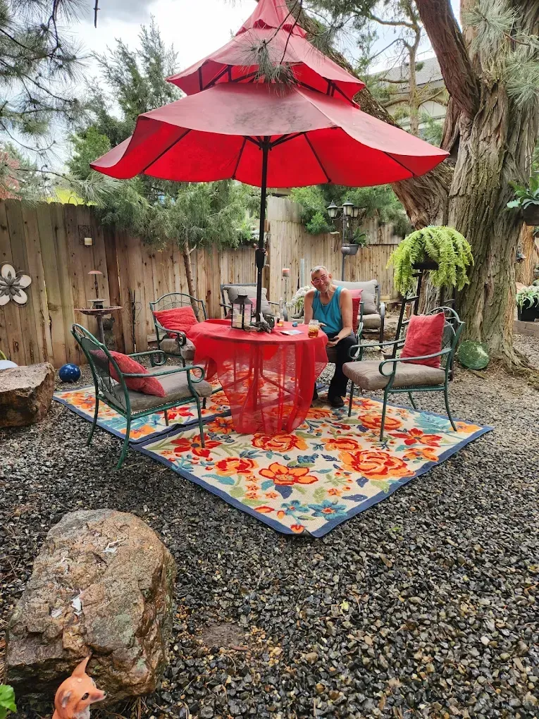 A woman sits at a table under a red umbrella in a backyard patio.