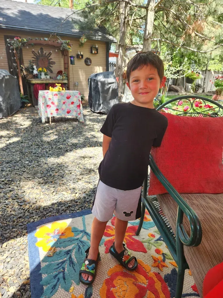 Boy in black shirt and shorts, smiles outdoors. Stands near a chair and patio with table.