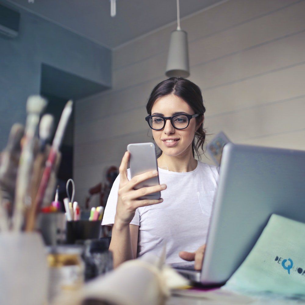 A woman wearing glasses is using a laptop while holding a cell phone