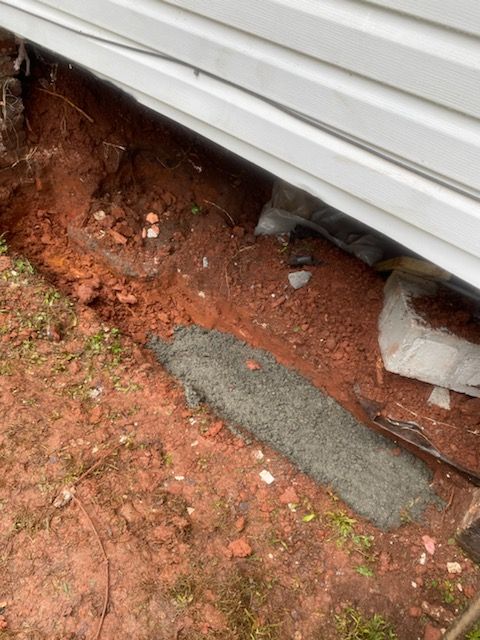 Dug trench along the side of a white house. Red dirt with a gray concrete piece lying in it.