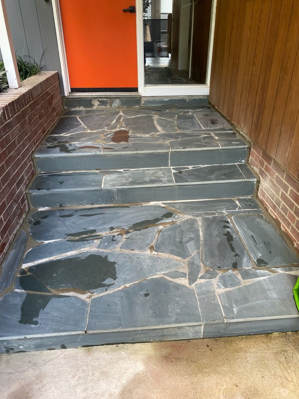 Outdoor steps leading to a door with orange paint. Flagstone steps are dark grey. Brick and wood siding are visible.