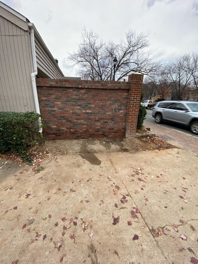 Brick wall next to a building, by a sidewalk and street; bare tree and cars in the background.
