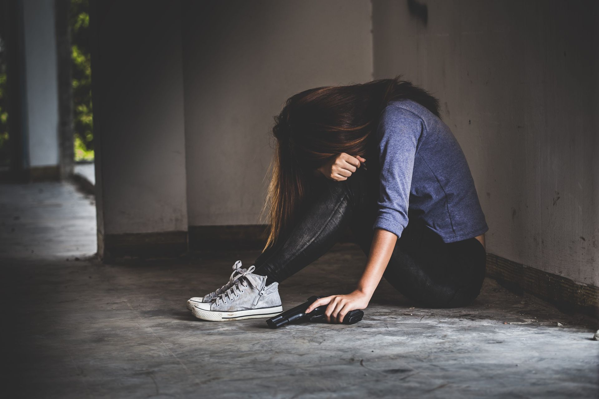 Person sitting on the floor in a dim corridor, head down beside white sneakers, appearing upset or exhausted