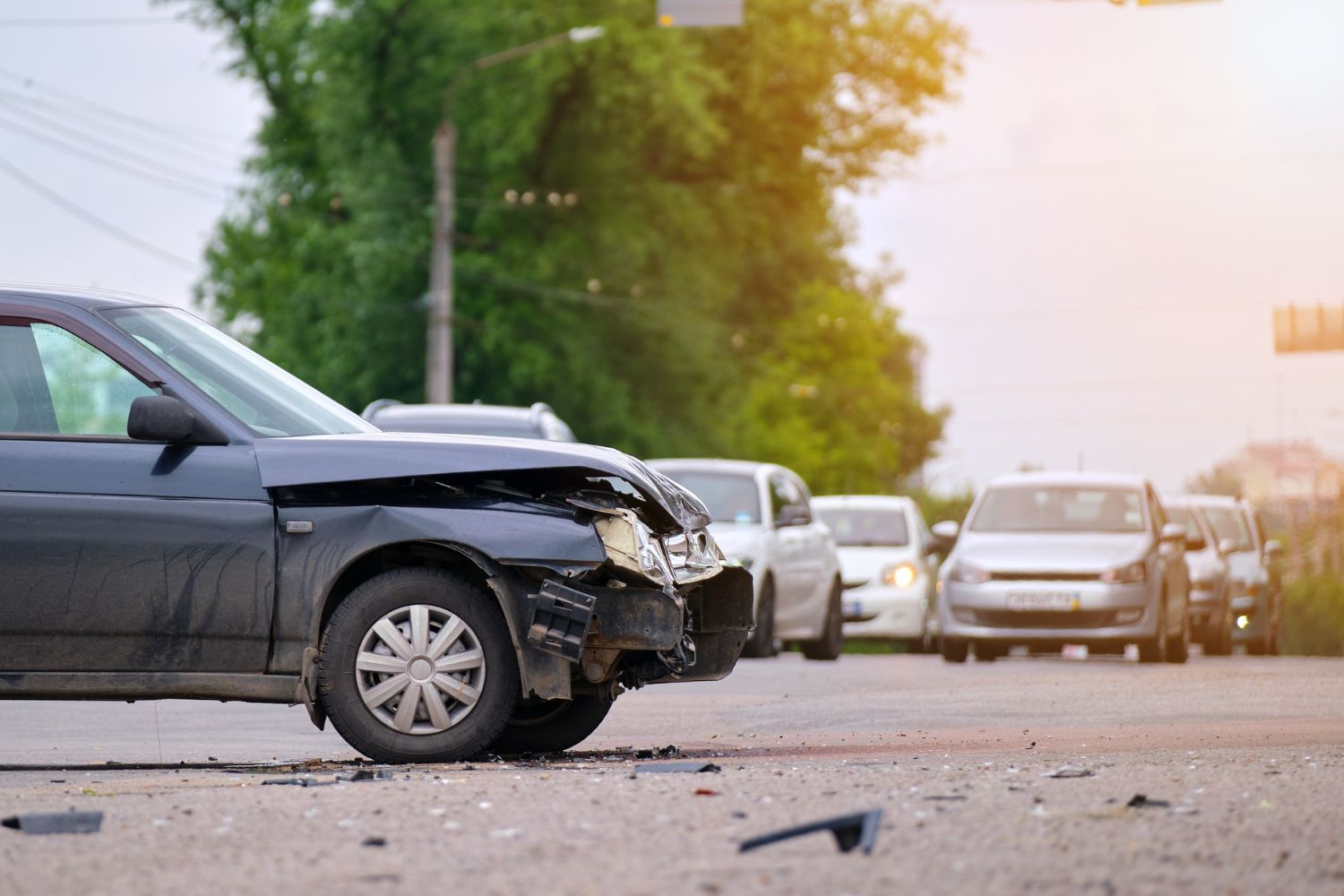 A dark-colored car with front-end collision damage parked on a road with other vehicles in the background.