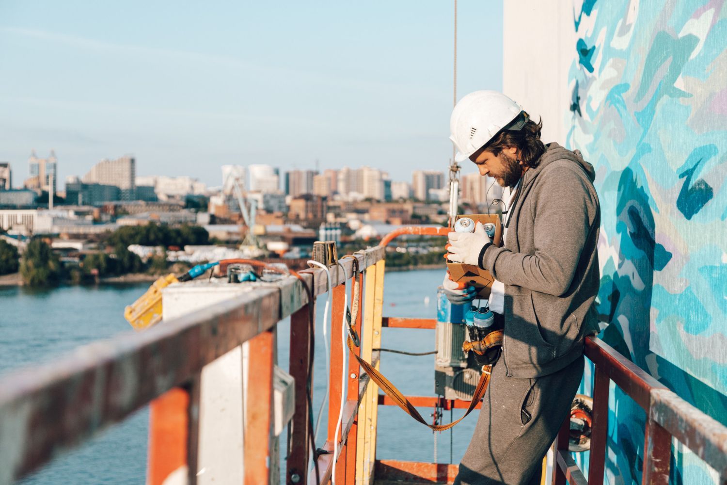 A person in a hard hat and grey clothing works on a wall mural from a scaffolding platform above a river and city.