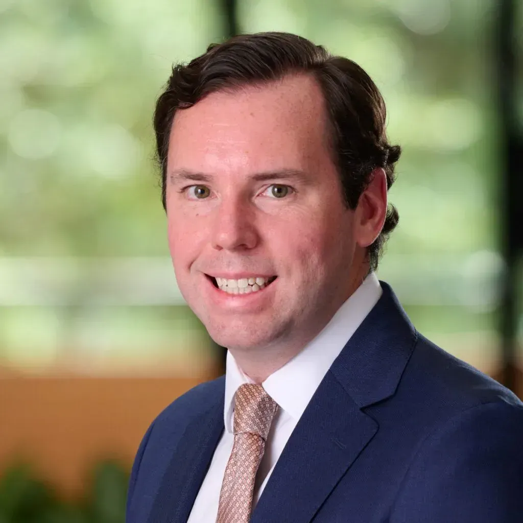 Headshot of a smiling person wearing a blue blazer, white collared shirt, and patterned tie against a soft, green background.