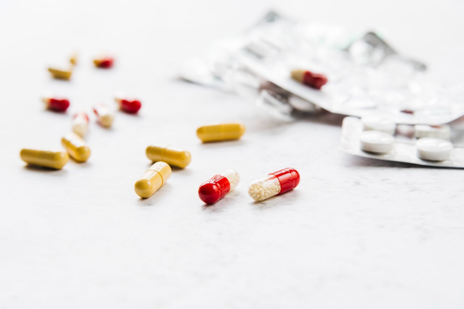 A collection of yellow and red-and-white capsules scattered on a white surface, with empty blister packs in the background.