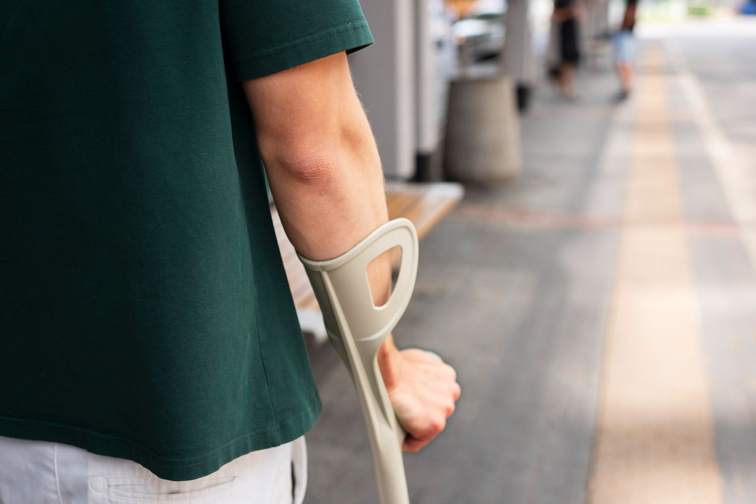 A person in a green shirt holds a beige forearm crutch while standing on a walkway.