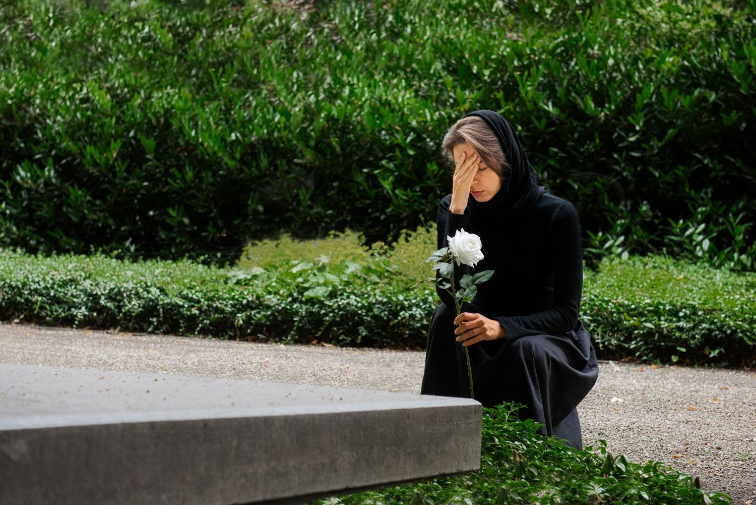 A person in black clothing kneels by a grave, covering their face with one hand while holding a white rose in the other.