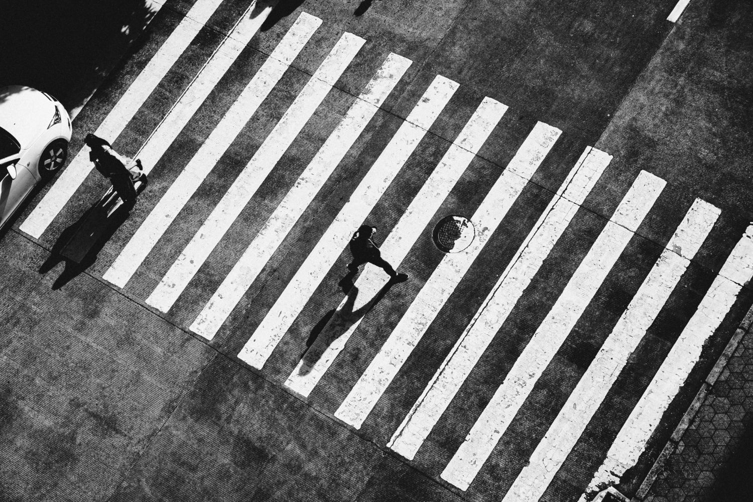 A high-angle, black-and-white photo of two people walking across a striped pedestrian crosswalk next to a car.