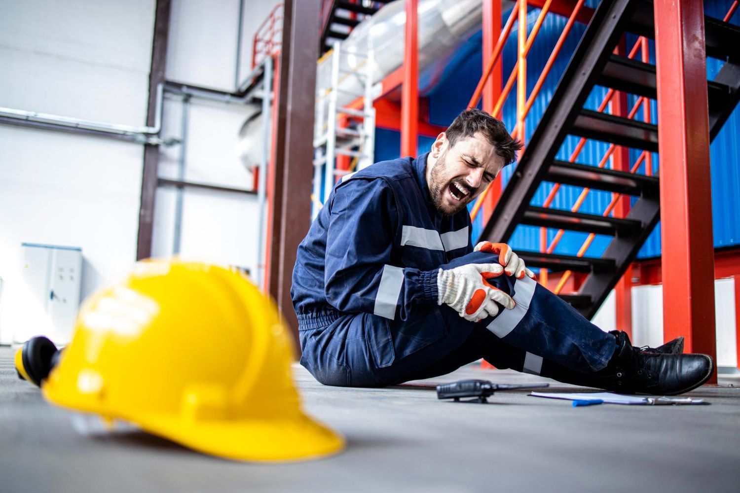 A construction worker in a blue uniform sits on an industrial floor, clutching an injured knee in pain.