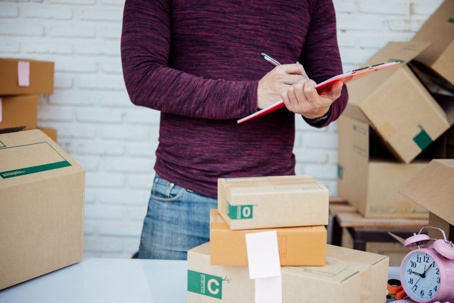 A person in a maroon sweater stands among cardboard shipping boxes, writing on a red clipboard.