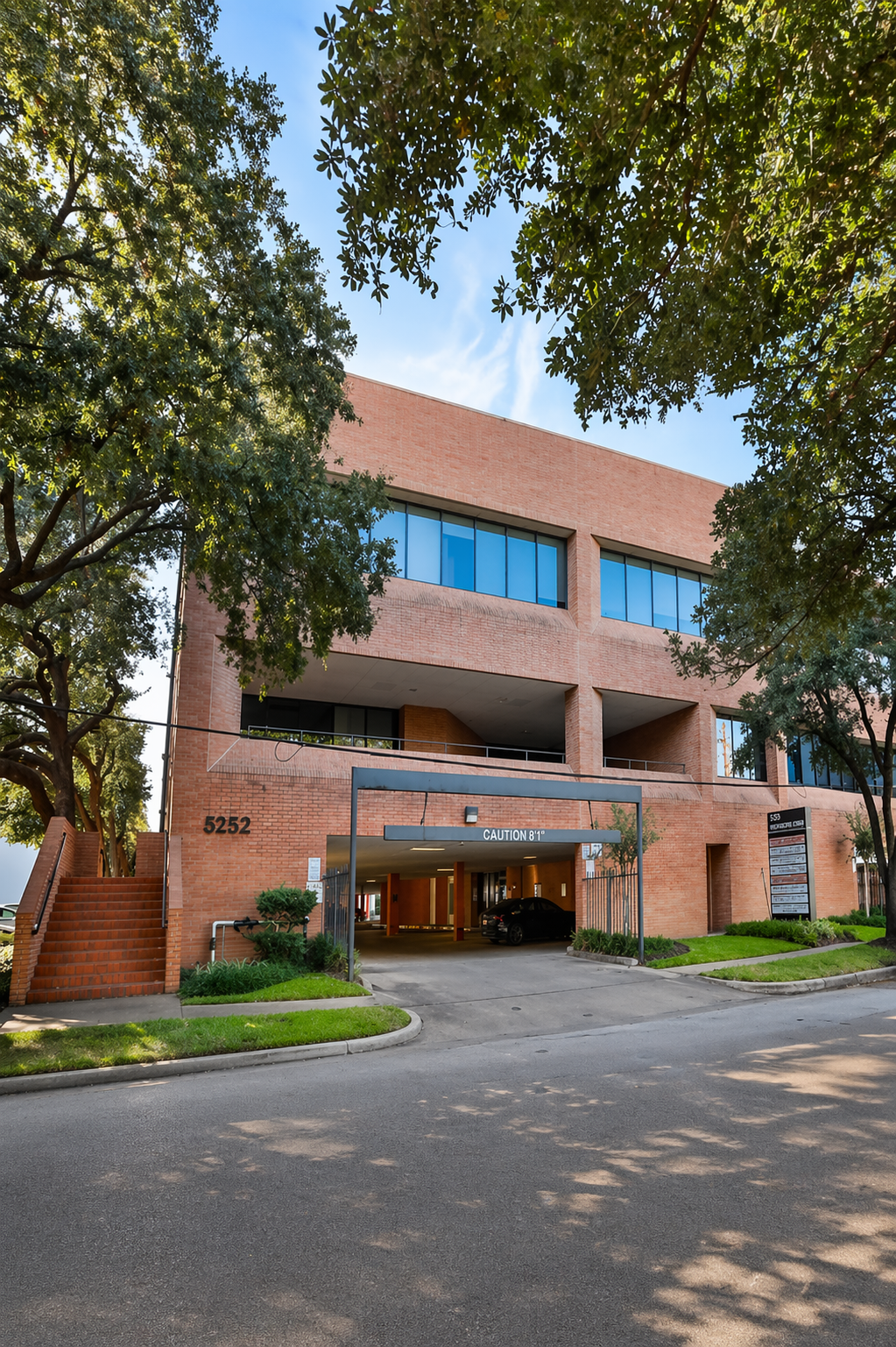 A paved path lined with large, leafy trees leads toward 5252 Westchester Street building surrounded by greenery and shrubbery.