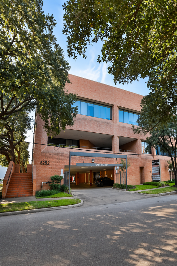 A paved path lined with large, leafy trees leads toward 5252 Westchester Street building surrounded by greenery and shrubbery.