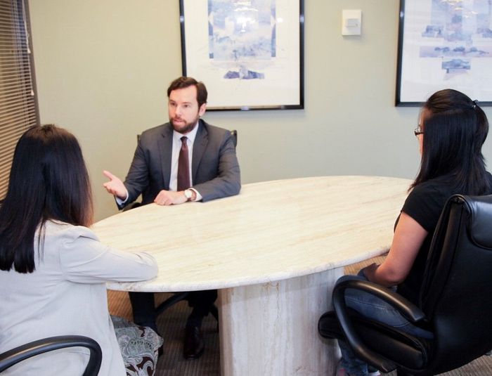 A professional in a suit gestures while sitting at an oval table with two others in a lit office with framed art.