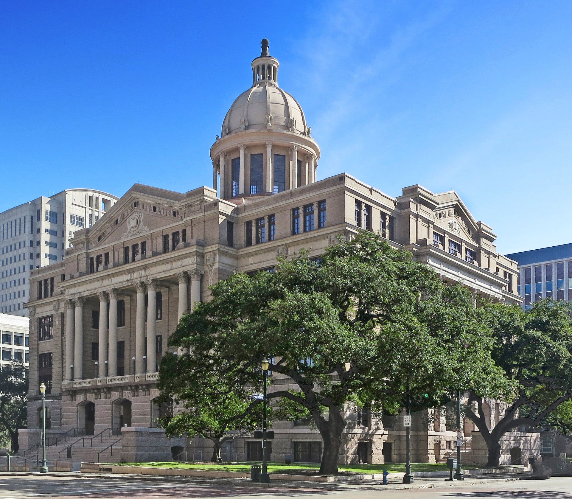 A grand, light-colored stone courthouse with a central dome, framed by large green trees under a clear blue sky.