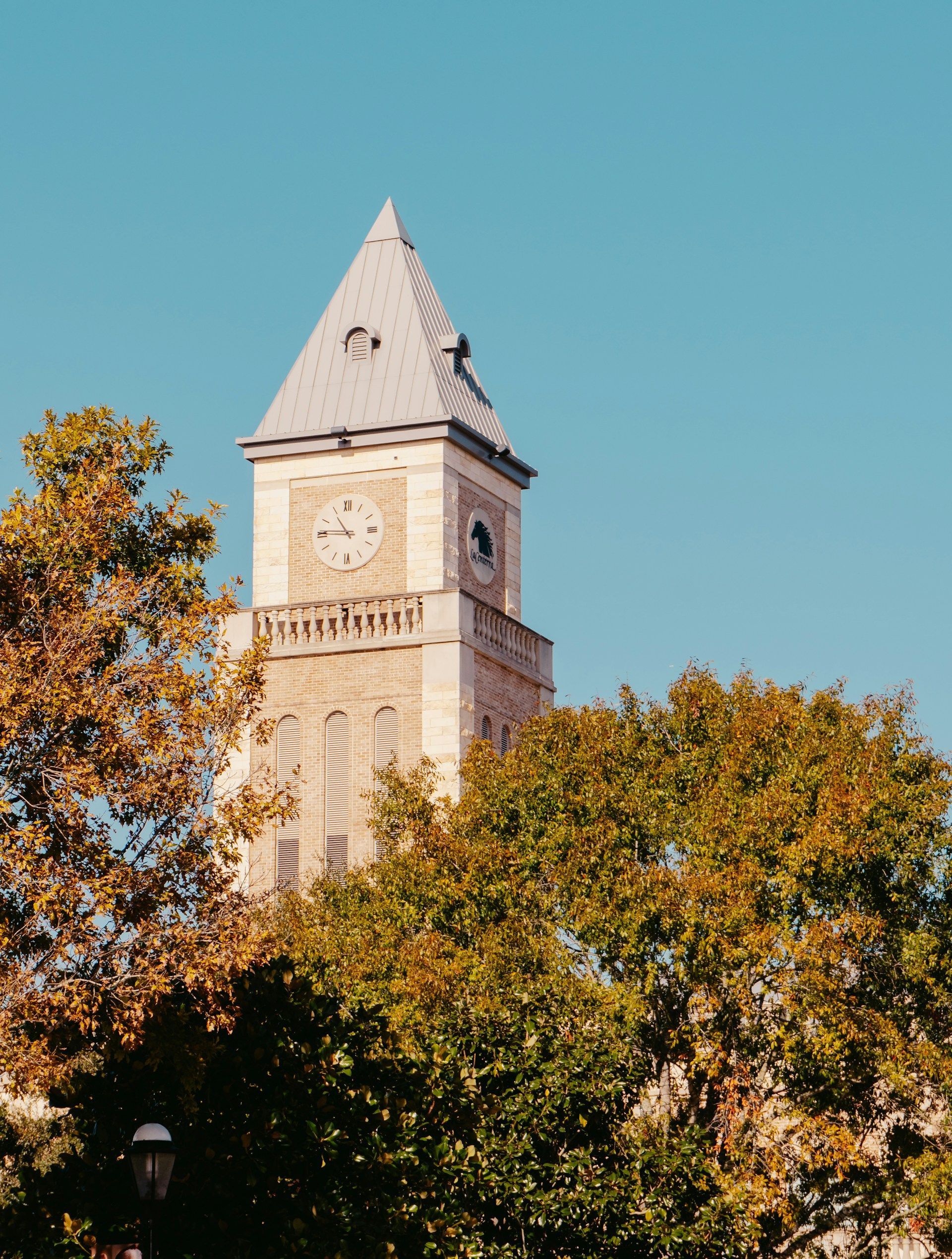 A stone clock tower rises behind autumn trees against a clear blue sky.