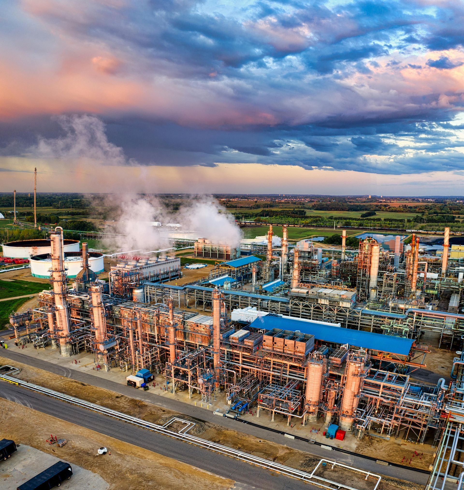 An aerial view of an industrial oil refinery at sunset, featuring tall pipes, storage tanks, and steam rising into clouds.