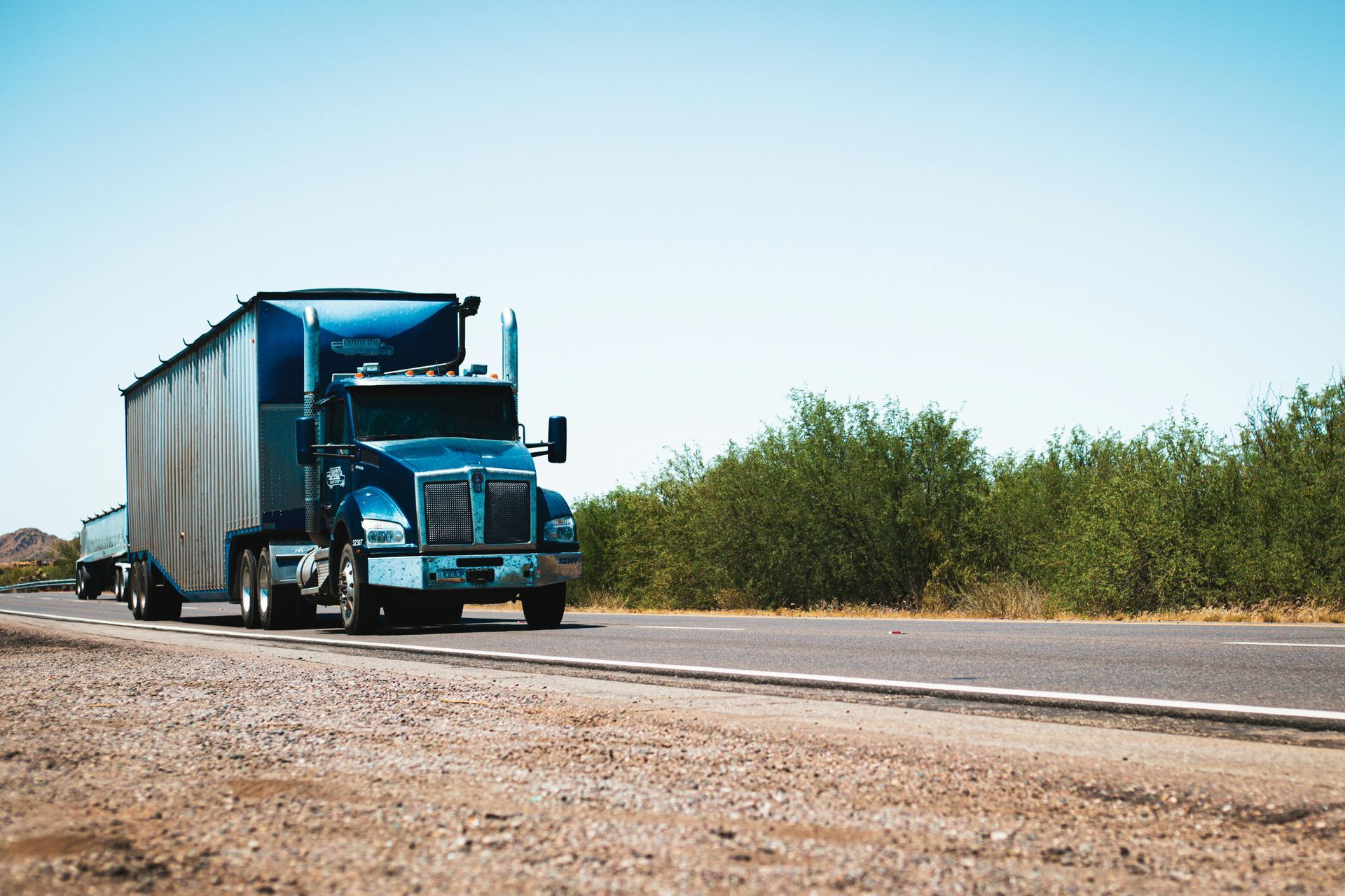 A blue semi-truck with a silver trailer drives along a paved road next to green trees under a bright blue sky.