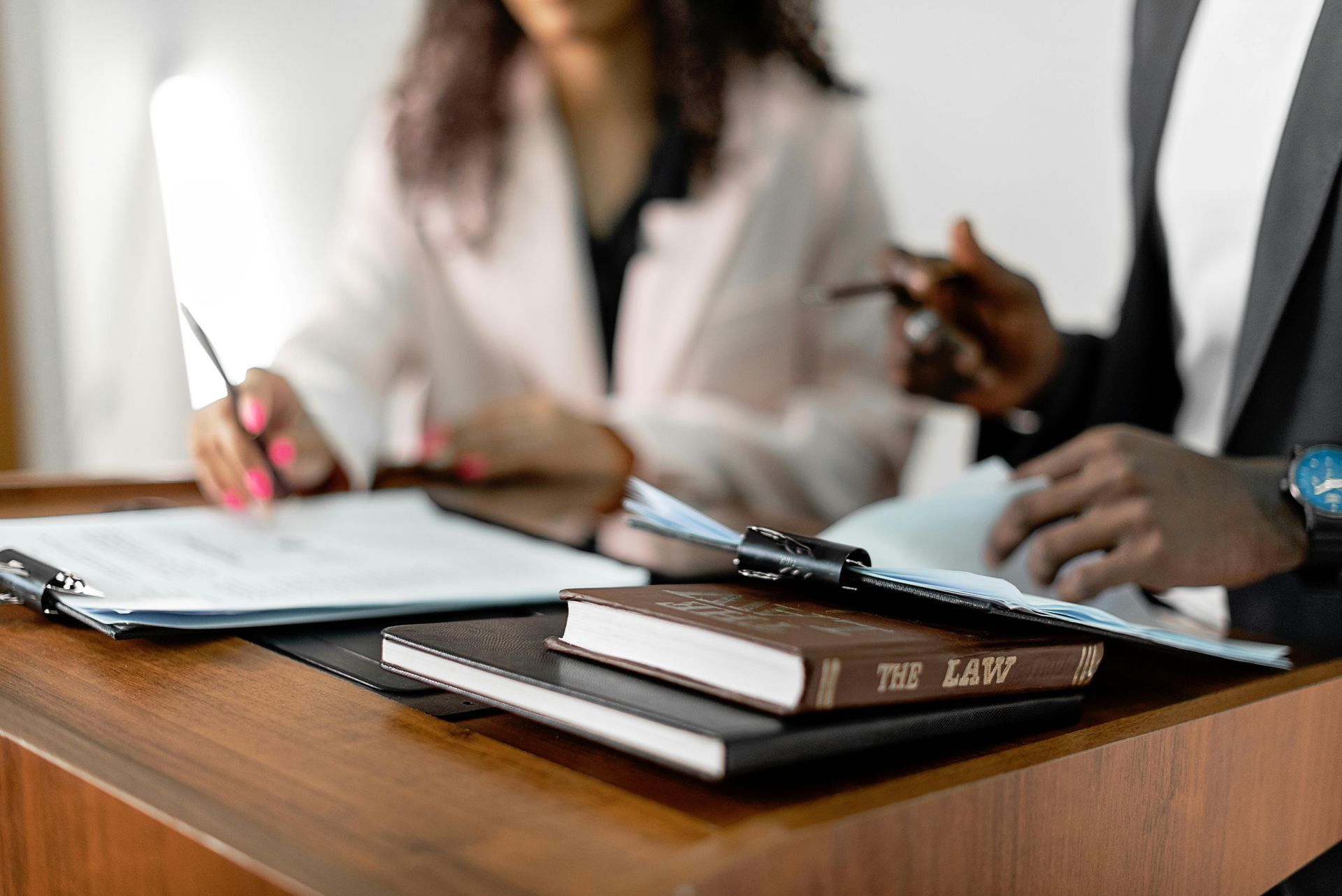 Two professionals in professional attire work together at a desk, reviewing documents and a book titled 