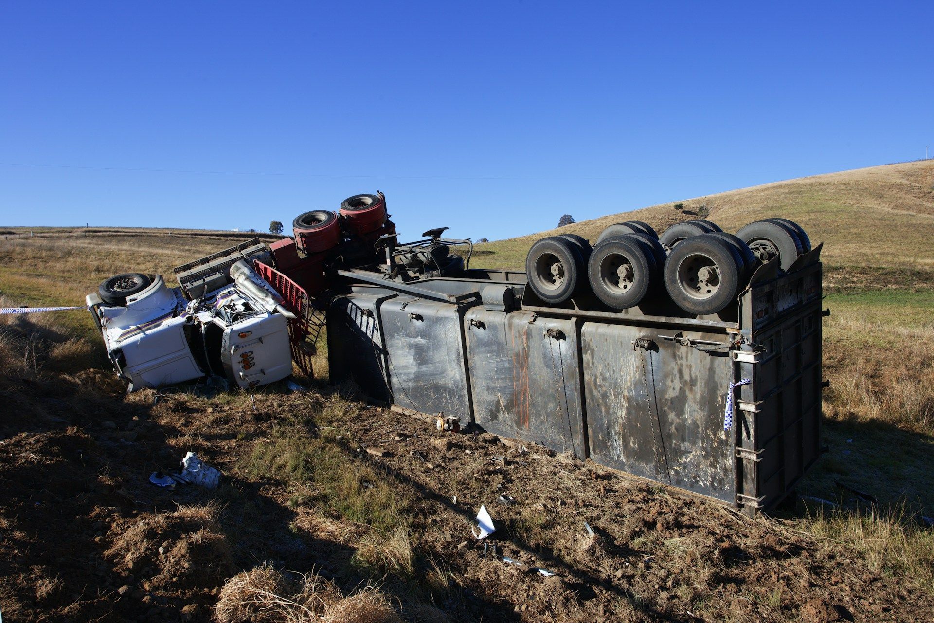 A white semi-truck lies on its side in a rural, grassy field under a clear blue sky.