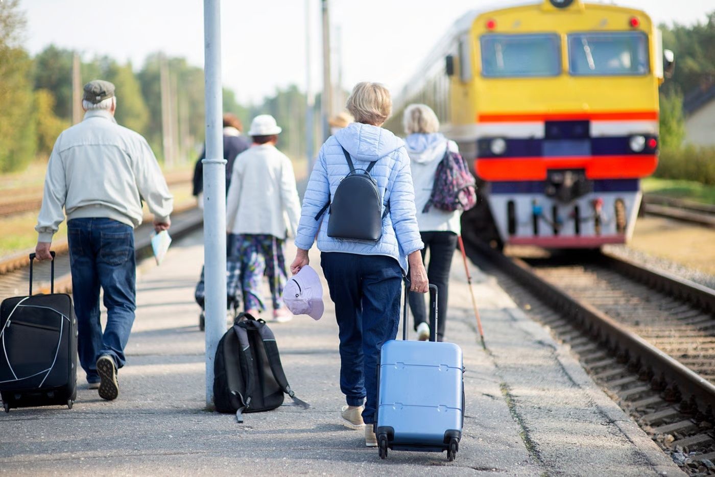 A group of travelers walks along a train platform toward an arriving yellow and white locomotive.