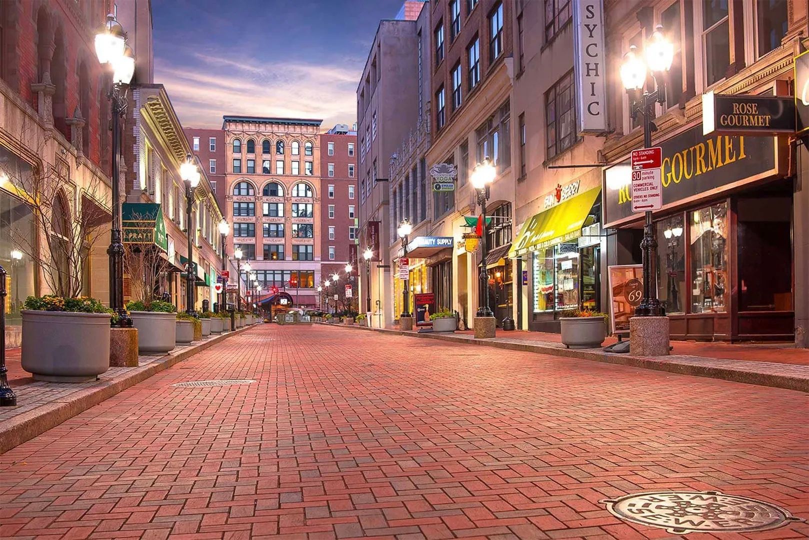 Cobblestone street lined with brick buildings, streetlights, and storefronts at dusk.