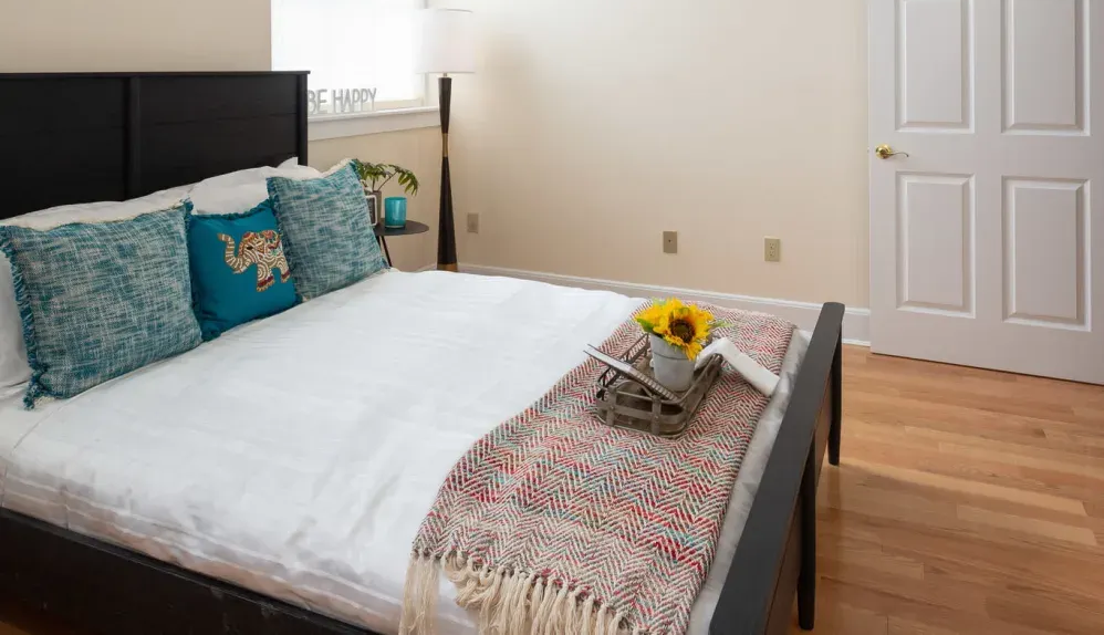 Bedroom with a black bed frame, white bedding, teal pillows, and a blanket; a tray with sunflowers sits on the bed.