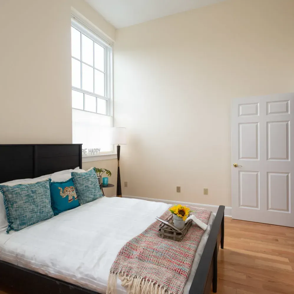 Bedroom with a bed, window, door, and beige walls. Light blue pillows, a blanket, and flowers are on the bed.