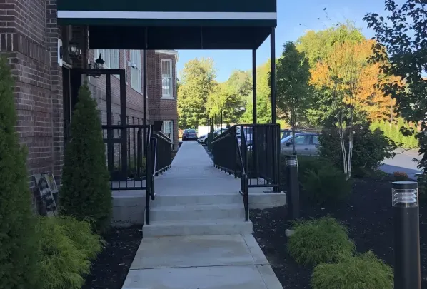 Brick building entrance with ramp, green awning, black railings, and trees on a sunny day.
