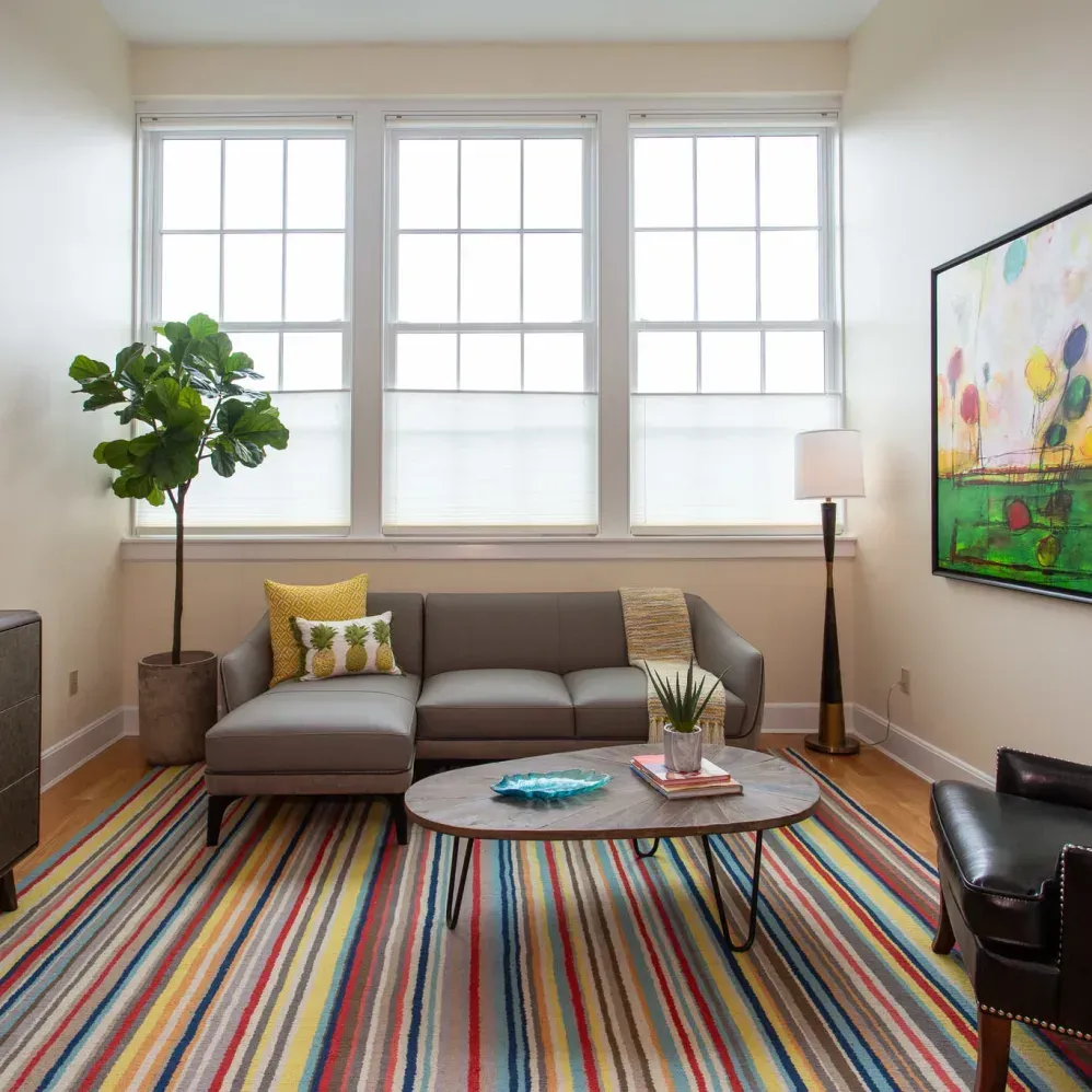 Living room: gray sofa, colorful striped rug, artwork, large windows, and a potted plant.