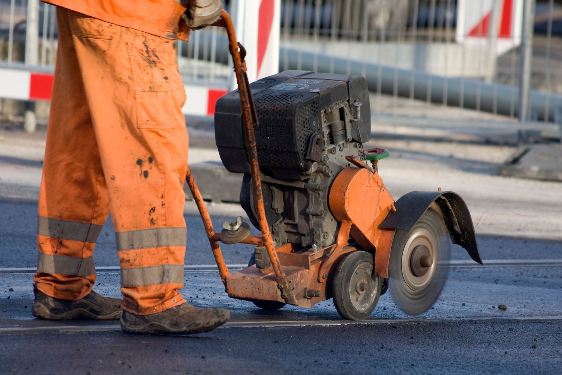 Worker in orange overalls using a concrete saw on asphalt.