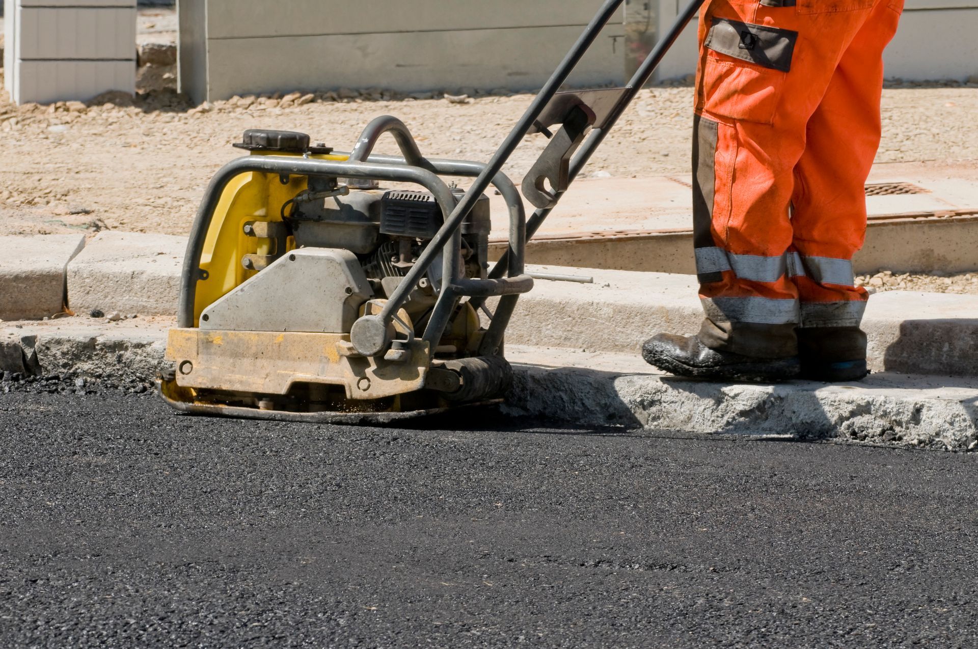 Person in orange work pants operating a plate compactor on fresh asphalt near a curb.