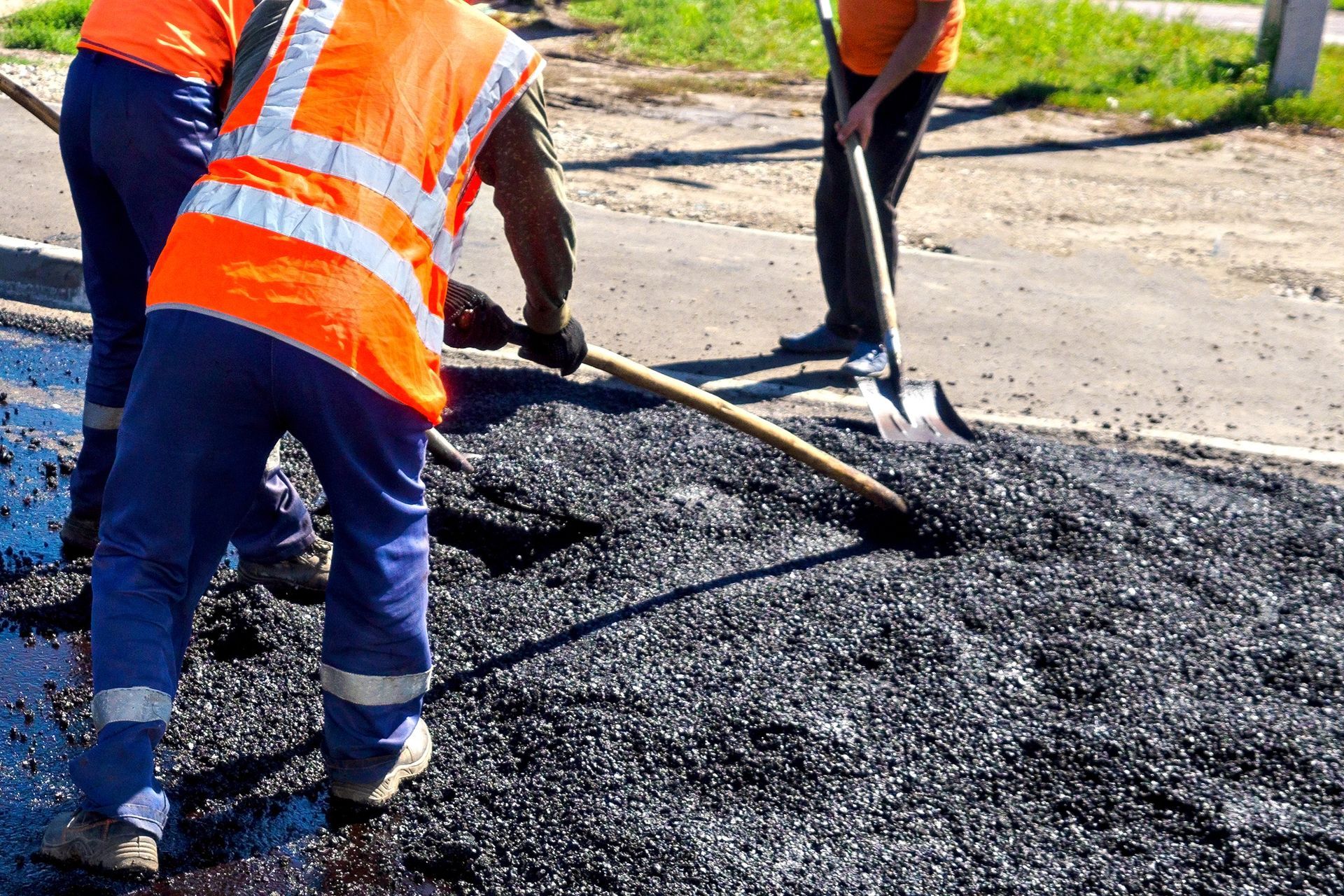 Workers in orange vests spreading asphalt on the road with shovels.