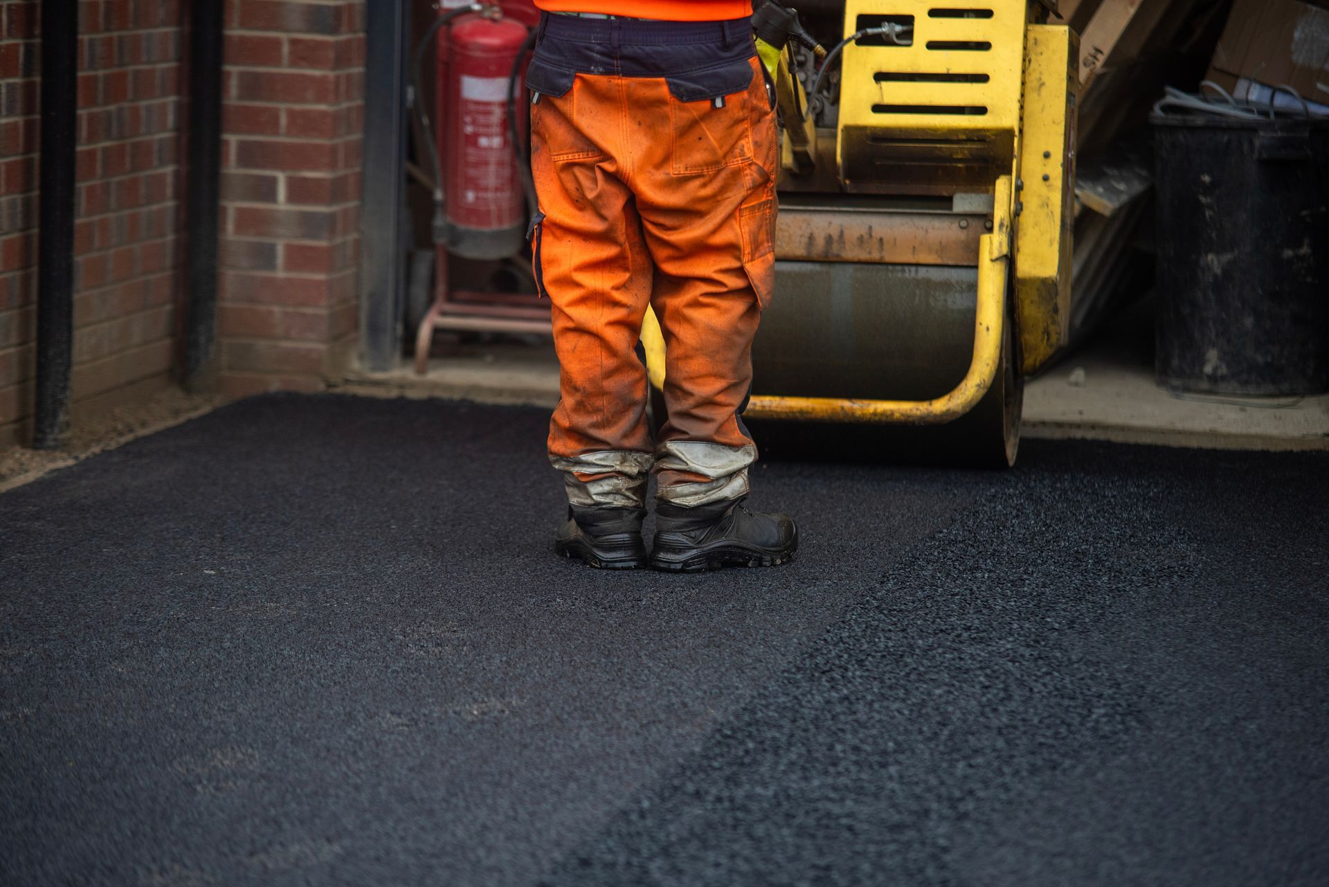 Worker in orange overalls stands on freshly laid black asphalt near a yellow road roller.