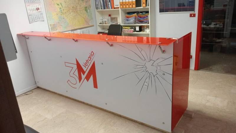 Banco della reception con marchio rosso e bianco, motivo in vetro crepato e logo aziendale.