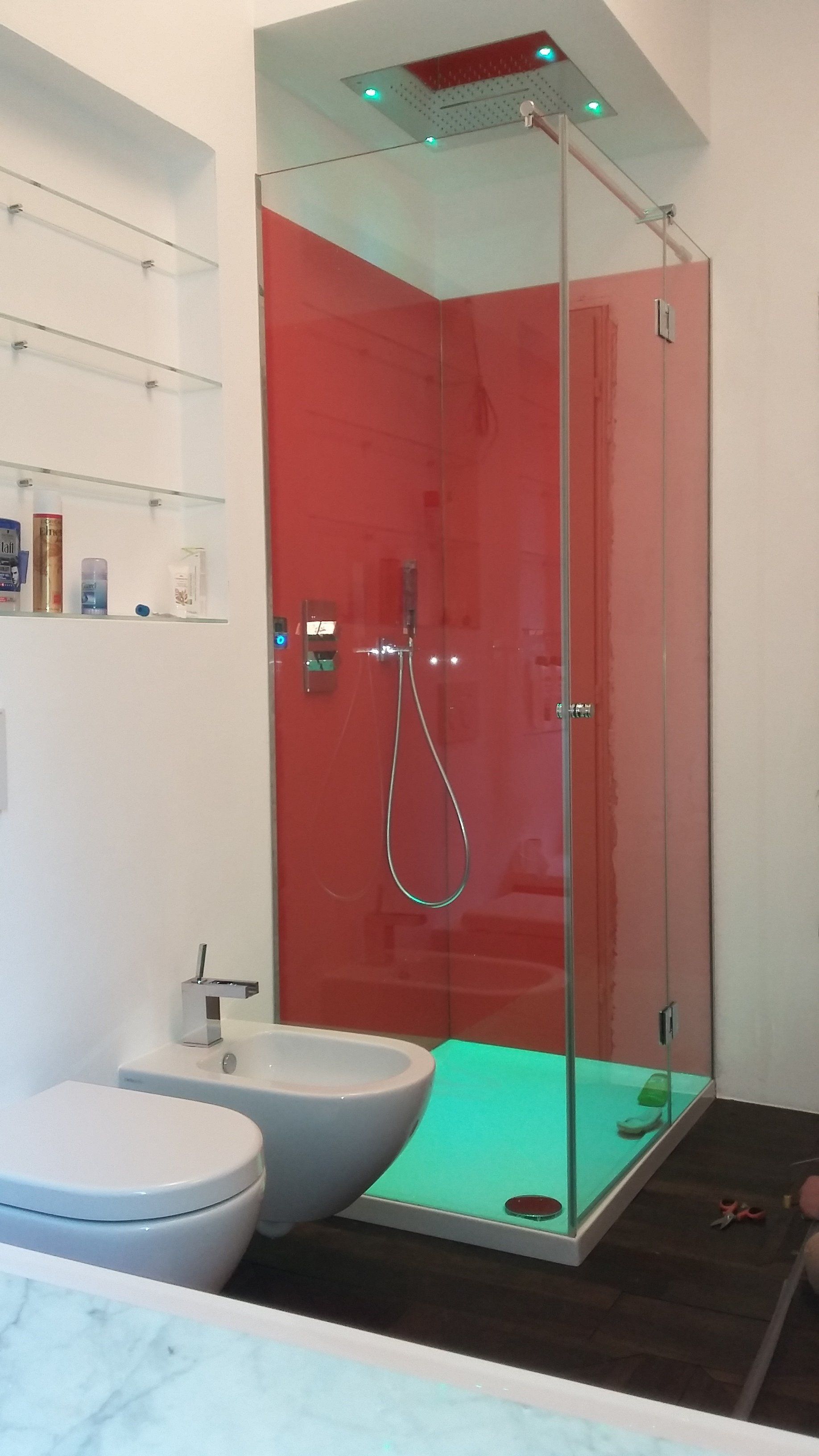 Bagno moderno con pannello doccia rosso, cabina in vetro, WC, bidet e illuminazione verde.