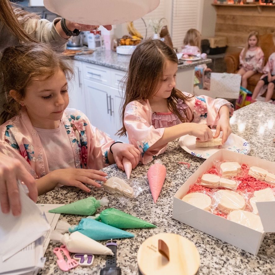 Two little girls are sitting at a table decorating a cake.