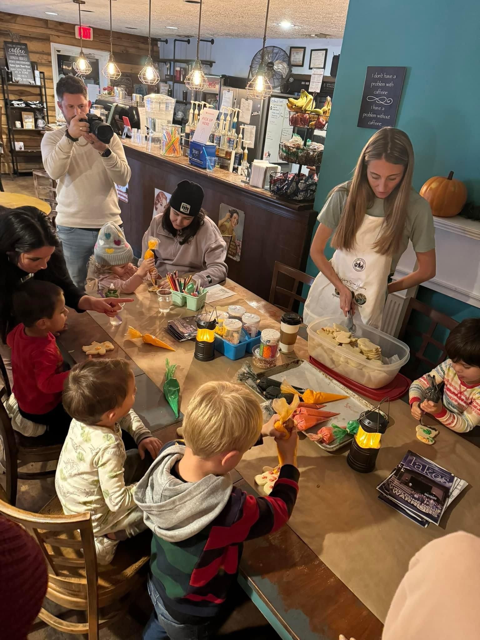Children decorate cookies with an instructor in a cafe. The instructor cuts cookies, and a person takes a photo.